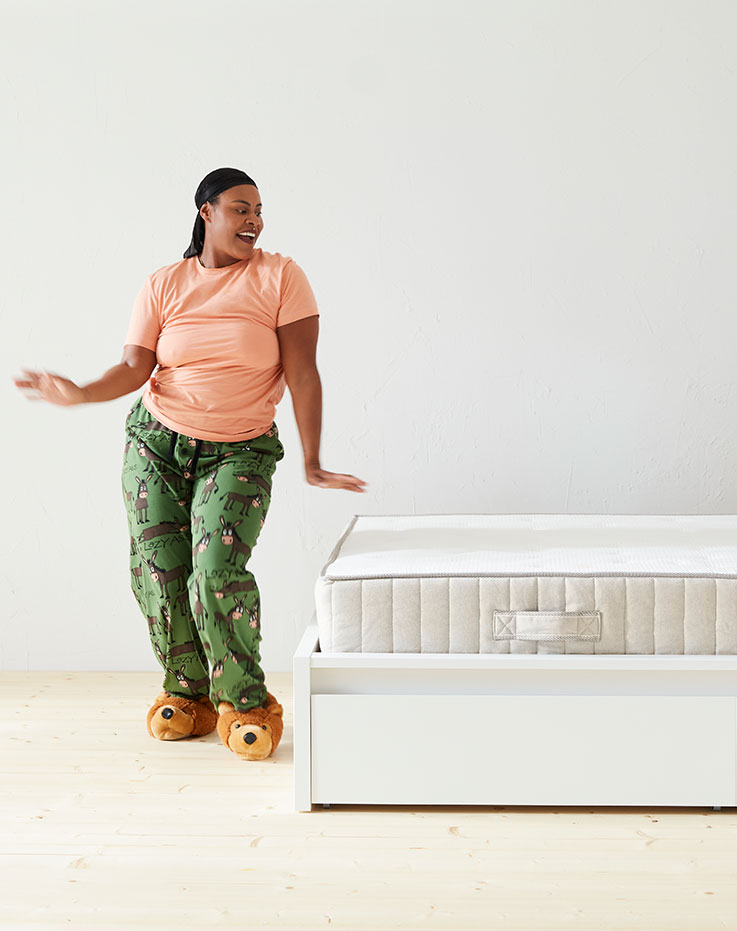 Mujer de pie en una pose dinámica delante de una cama con un colchón blanco visible, el plano está totalmente iluminado y blanco.