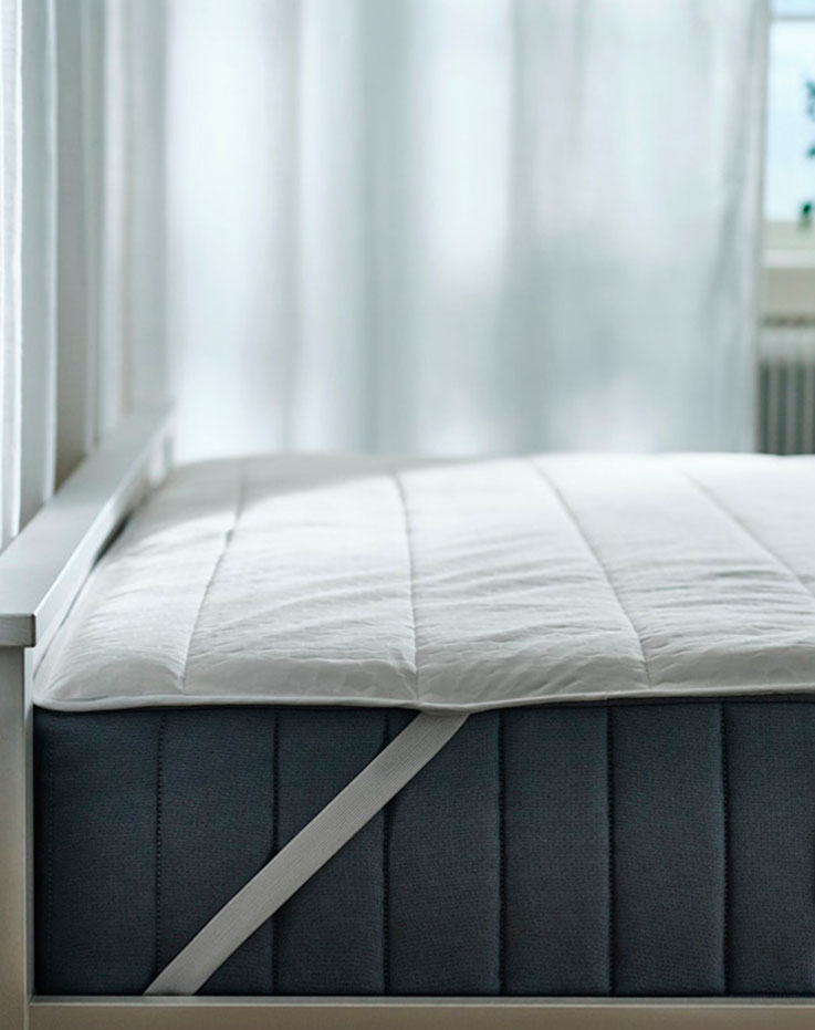 Imagen de una cama con un colchón gris visible y el protector de colchón con la cinta que lo sujeta.