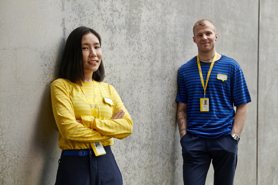 Two IKEA employees in yellow and blue uniforms posing in front of a gray concrete wall.
