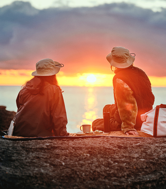 Two people wearing SOLUPPGÅNG hats are sitting on a SOLUPPGÅNG picnic blanket while watching the sunset by the sea.