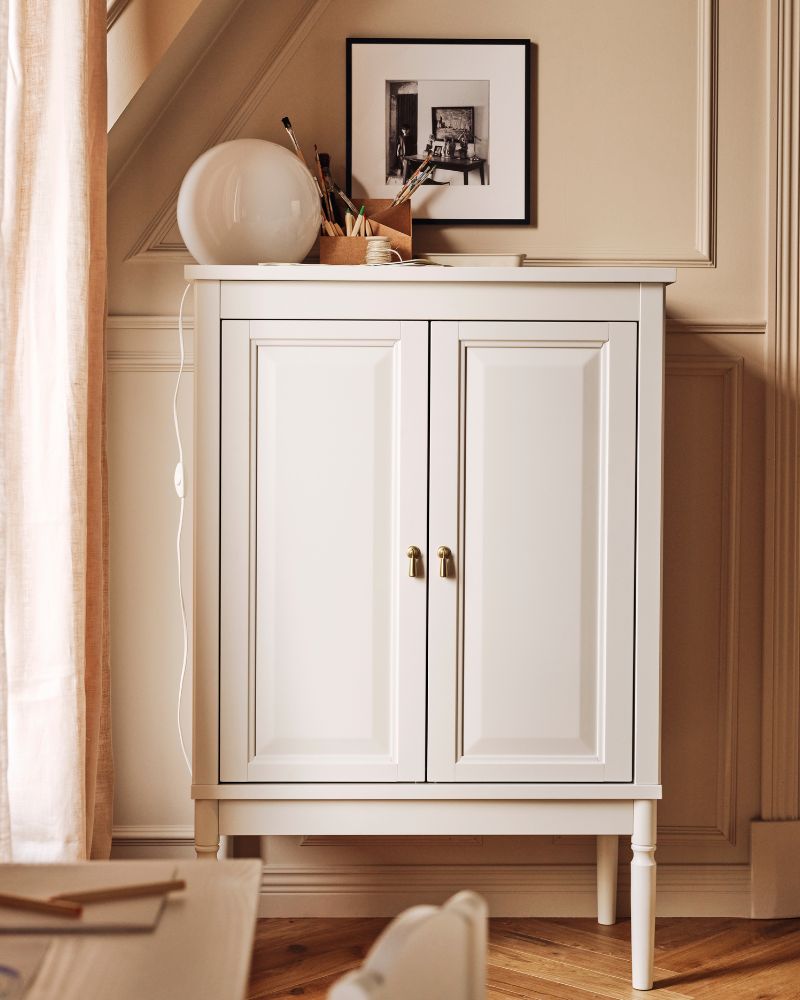 An off-white FÄRJKARL cabinet with a FADO table lamp on top stands against the wall near a window in a living room.
