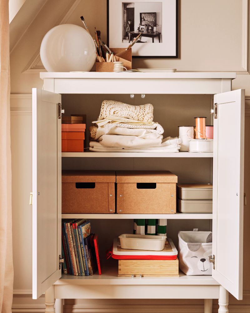 An off-white FÄRJKARL cabinet with many things on its shelves including two GÄRDESGÅRD storage boxes and a DRÖMSLOTT box.