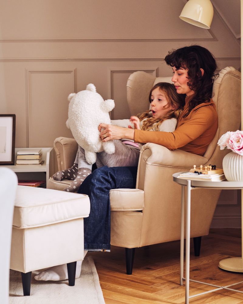 A child sits on an adult’s knee with a GREJSIMOJS soft toy on a STRANDMON wing chair. Beside them is a STRANDMON footstool.