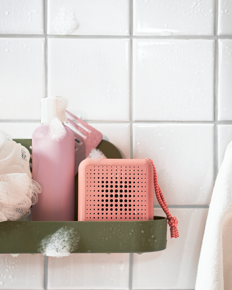 Estante de pared con ventosa verde grisaceo, con una esponja beige y un altavoz pórtatil impermeable rosa.