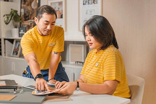 Dos empleados de IKEA con uniformes de rayas amarillas trabajan juntos en un escritorio, mirando un círculo cromático y muestras de materiales.
