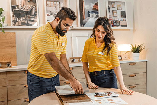 Dos empleados de IKEA con camisetas polo de rayas amarillas están junto a una mesa revisando documentos de diseño y muestras de materiales para encimeras en una sala de exhibición.