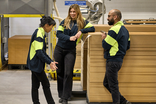 Tres trabajadores de logística con chaquetas oscuras y paneles amarillos de alta visibilidad conversan y ríen junto a grandes pilas de cajas de cartón embaladas en plano en un almacén.