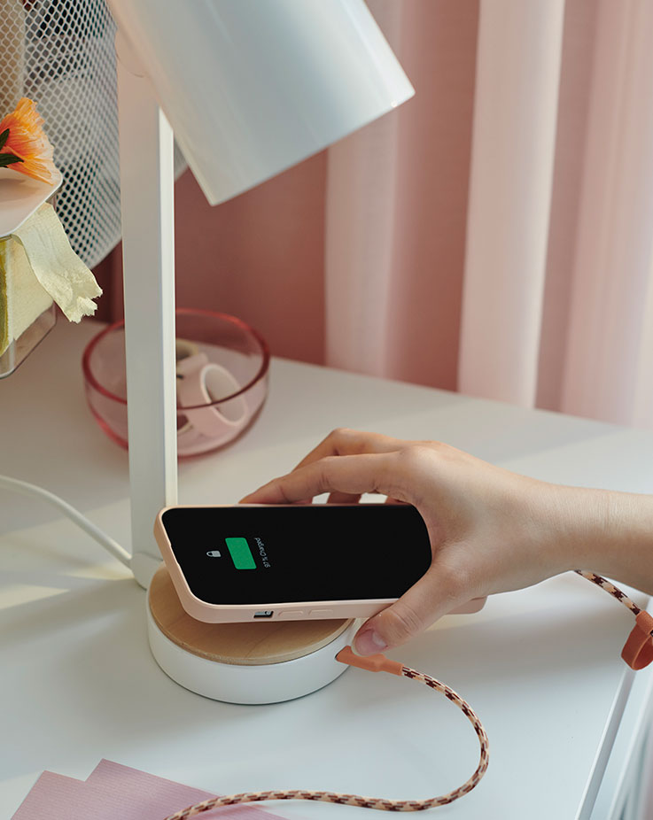 A hand holds a mobile phone connected by a cable to a USB-C port in the foot of an ENSMÄRKE desk lamp.