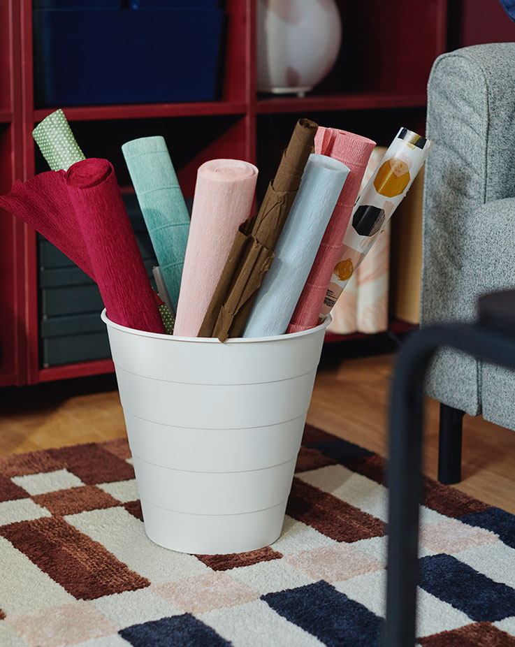 A white FNISS waste bin with rolls of coloured paper in it stands on a MARKBLÅMME rug in front of red shelving units.