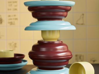 A SPJUTLILJA 3-piece serving set with dark purple bowls, yellow cups and light blue plates, in a tower on a dark table.