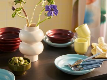 A dining table set with a SPJUTLILJA 3-piece serving set, including a dark purple bowl, a yellow cup and a light blue plate.