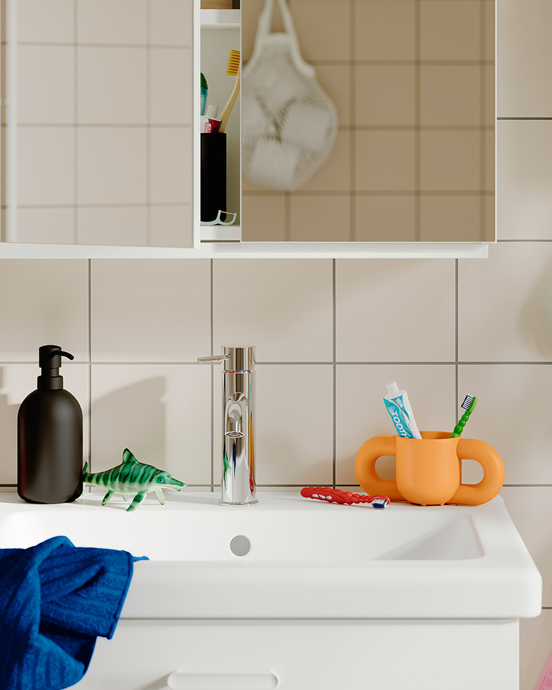 An orange HUMLESJÖN mug containing toothbrushes on a bathroom counter beside a sink and soap dispenser.