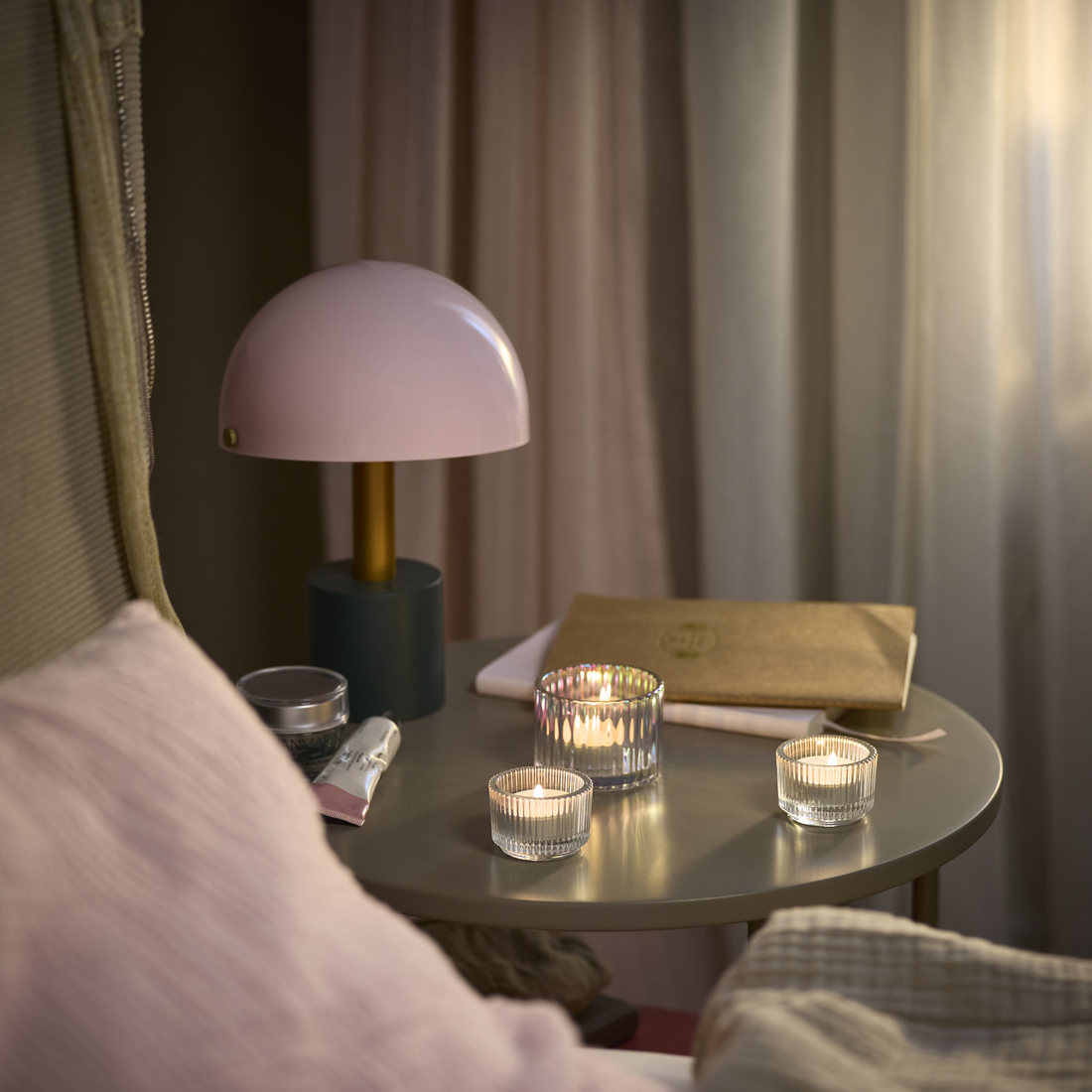 Mesa de noche con lámpara rosa y velas encendidas en portavelas de vidrio estriado. Ambiente íntimo y acogedor.