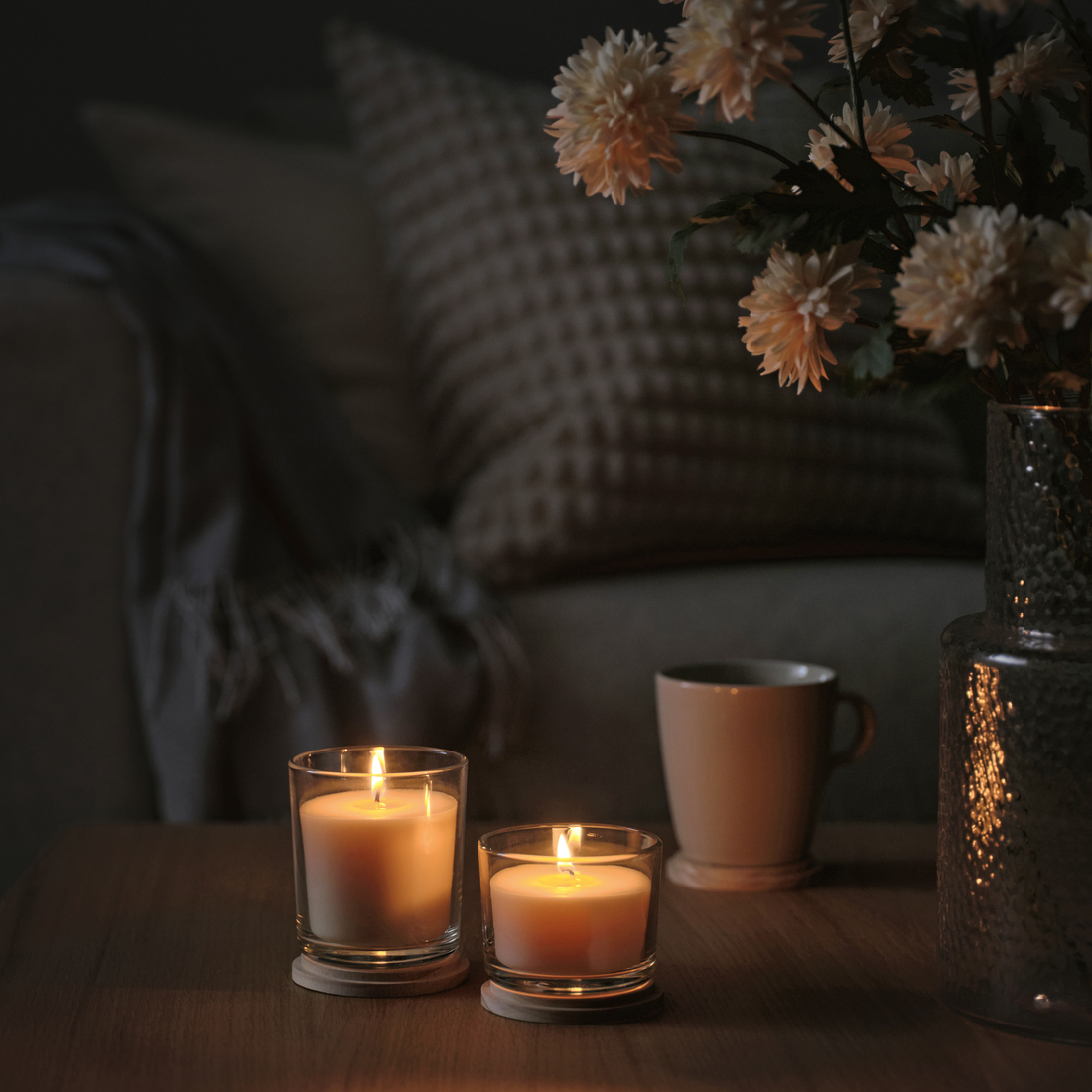 Dos velas encendidas sobre una mesa de madera, junto a una taza y flores. Ambiente hogareño, cálido y relajante.
