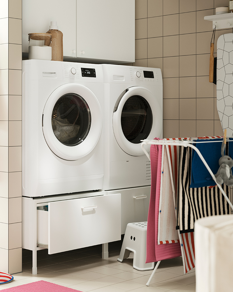 An ENHET pedestal, placed under a washing machine and drier, in a tiled laundry room with a drying rack.