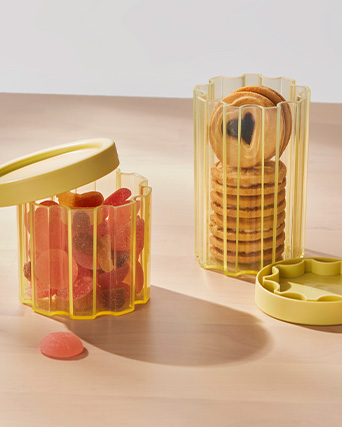 Frascos acanalados amarillos con galletas y gominolas sobre una mesa de madera con iluminación brillante y cálida.
