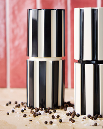 Dos molinillos de especias con rayas blancas y negras sobre madera, frente a una pared de azulejos rojos.