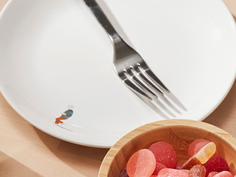 Plato blanco con tenedor metálico y un pequeño dibujo, junto a un cuenco de madera lleno de gominolas rojas.