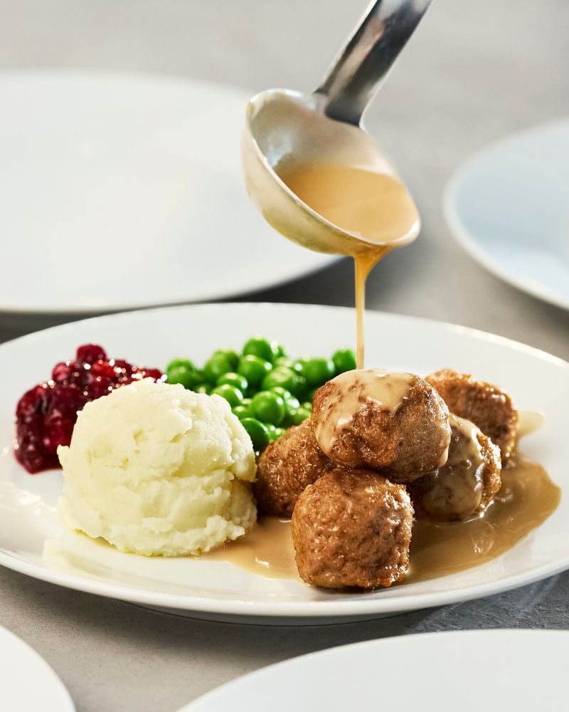Close-up of a plate of traditional meatballs with mashed potatoes, peas, and lingonberry jam. A spoonful of sauce pours creamy brown sauce over the meatballs, creating a continuous stream of moving sauce.