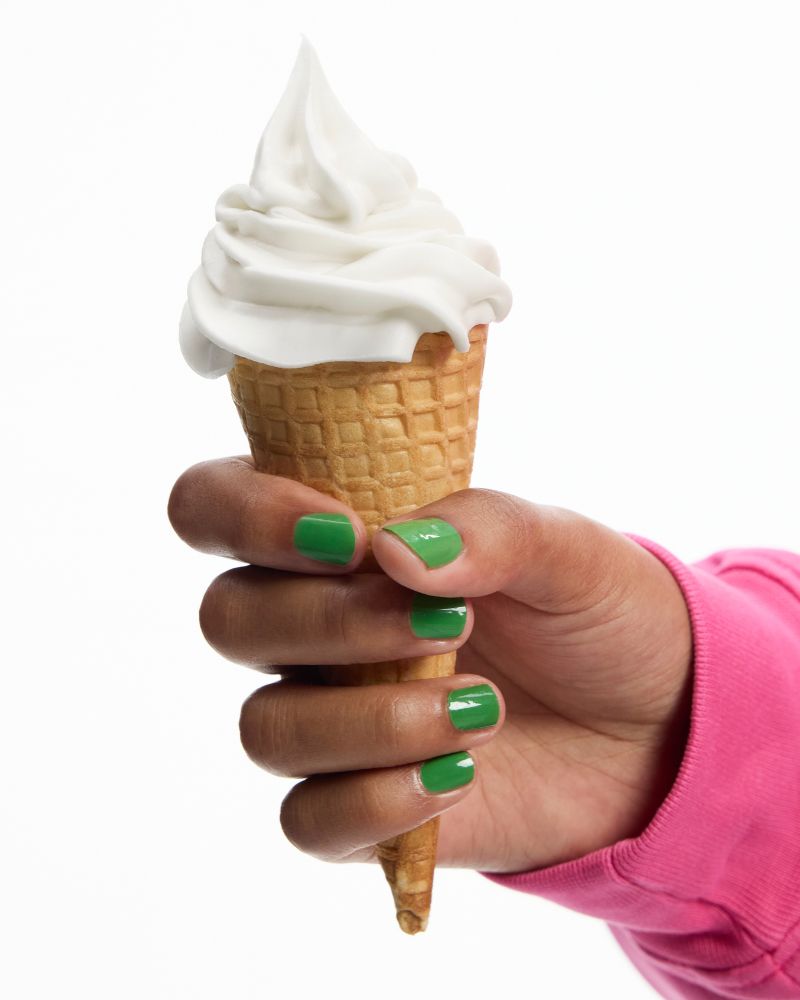 A hand with bright green nail polish holds a cone of soft-serve cream or vanilla ice cream against a clean white background. The person is wearing a vibrant pink sweatshirt.