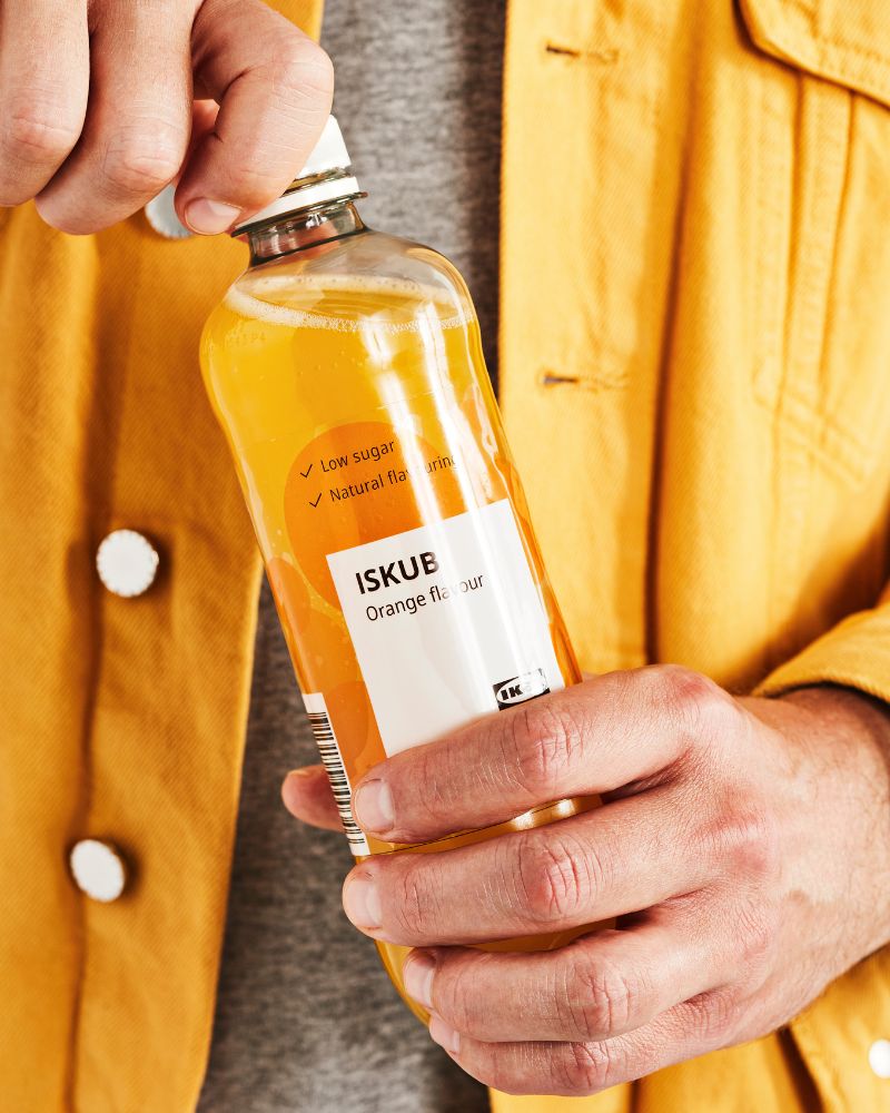 Close-up of hands opening a clear bottle of IKEA's ISKUB orange soda. The person is wearing a mustard-colored corduroy jacket and a gray T-shirt.