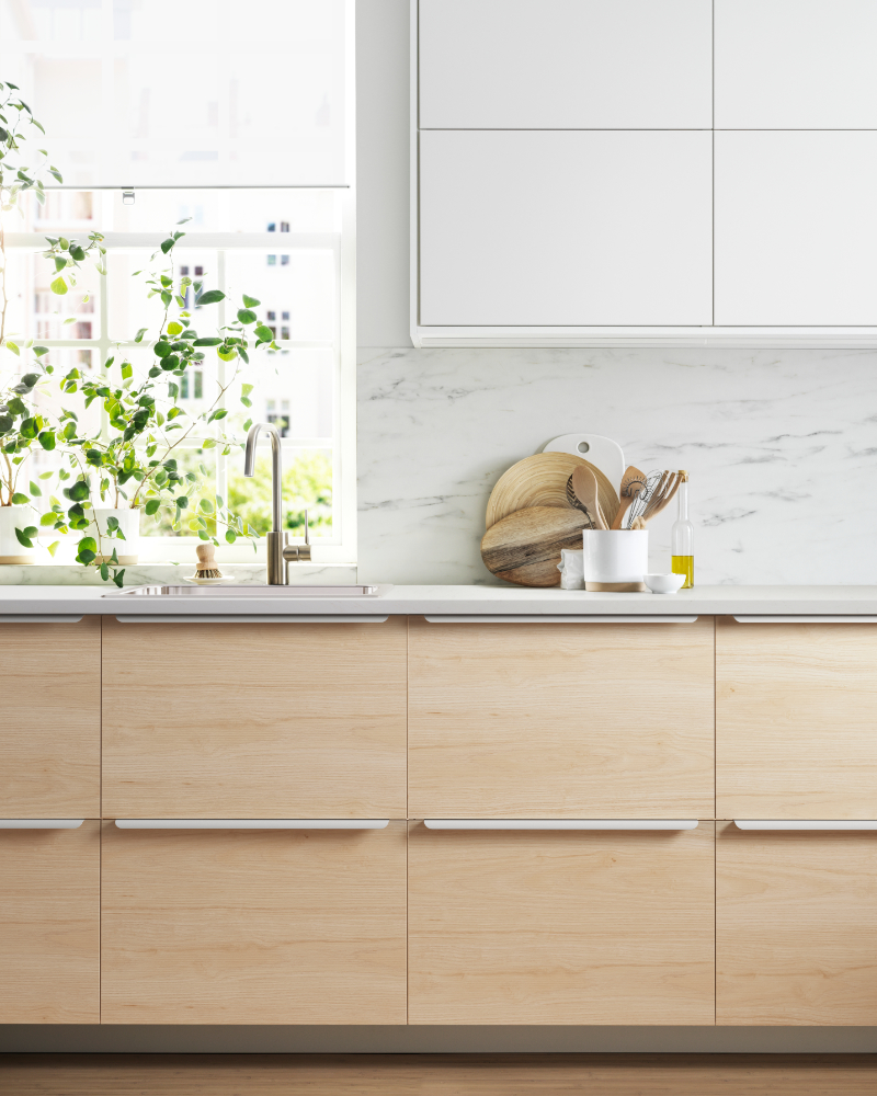 Cocina moderna y espaciosa con puerta a un jardín, los frentes de los cajones son de bambú, tiene colores blancos, se ven los hornos y la encimera de la cocina.