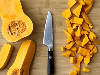 Vista superior de una calabaza violín cortada por la mitad, un cuchillo de chef con mango negro y dados de calabaza ordenados sobre una tabla de cortar de bambú.