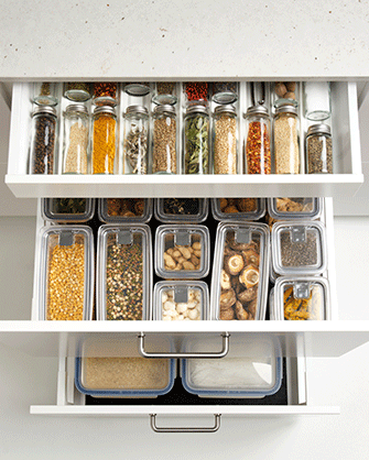 Vista superior de tres cajones blancos de cocina abiertos y organizados con tarros de cristal para especias y recipientes transparentes llenos de alimentos secos.