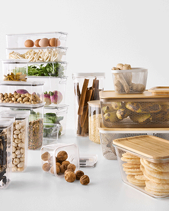 Un conjunto de recipientes de plástico transparente con tapa de bambú y cestas de rejilla ordenados sobre fondo blanco con ingredientes frescos y aperitivos.