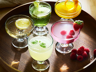 Cinco elegantes copas de cristal con cócteles de colores amarillo, verde y rosa decorados con frutas, dispuestas en una bandeja redonda de madera oscura.