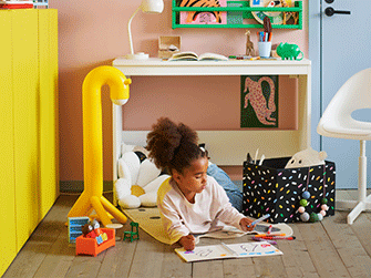 A child draws on the floor of a colorful room featuring a yellow giraffe-shaped lamp, a white daisy cushion, and a black storage box.