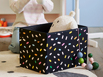 A black fabric storage box with a colorful sprinkle pattern holds a soft plush toy on a dotted rug.