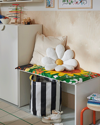 A white daisy cushion sits on a white bench with a colorful floral cover, next to a black and white striped storage basket.