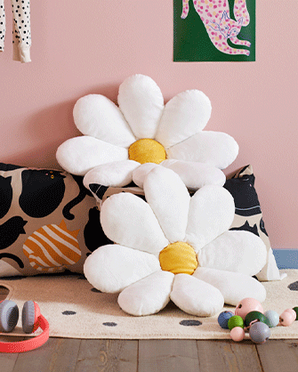 Two white daisy-shaped cushions with yellow centers rest on a cat-patterned pillow in a children's room with pink walls.