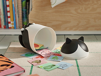 A cat-shaped white and black container tipped over on a checkered rug, with small illustrated cards spilling out.