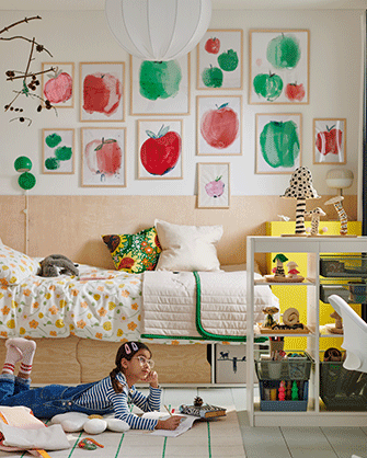 A children's bedroom featuring a wall decorated with framed apple prints, a bed with floral sheets, and a child drawing on the floor.