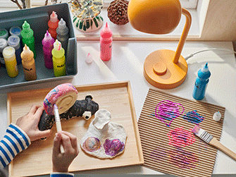 Overhead view of a child's hands painting clay figures on a wooden tray, next to a yellow desk lamp and colorful paint bottles.
