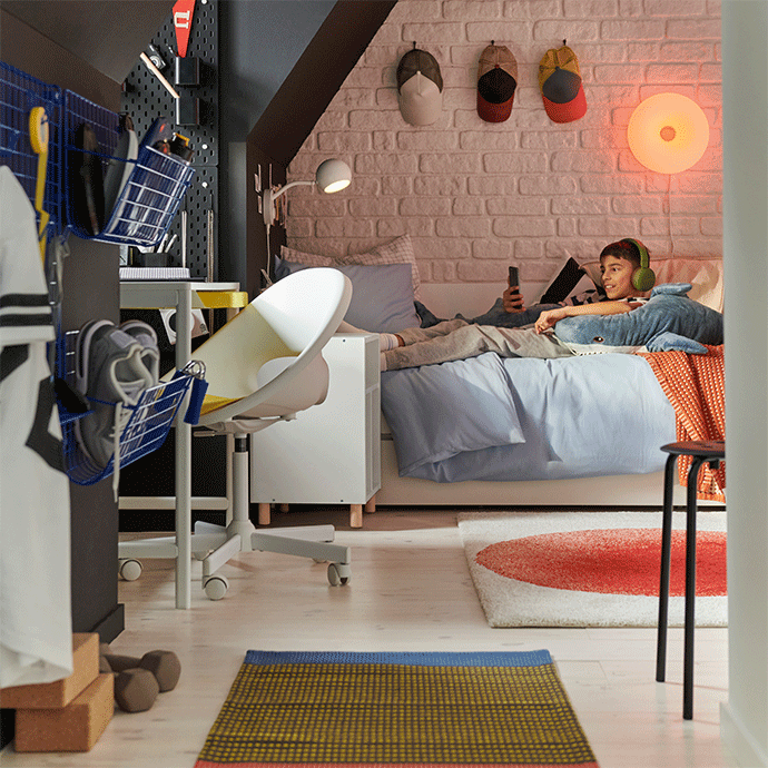 View of a modern youth bedroom under an attic slope, showing a desk with a white chair and a boy relaxing on a bed by a brick wall.