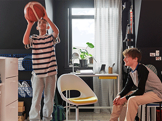 One boy prepares to shoot a basketball while another watches from a stool in a bedroom with a white desk and a swivel chair.