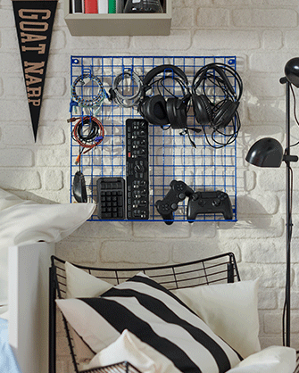 A blue wall-mounted wire grid panel holding headphones, cables, and gaming controllers above a black and white striped pillow.