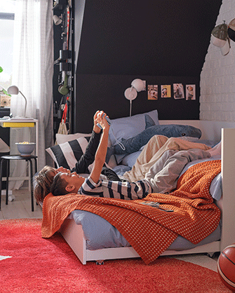 Two boys relax on a white bed with blue bedding and an orange textured blanket, one using a phone, in a bedroom with a red rug.
