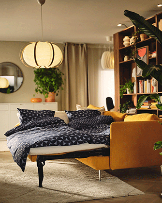 Sofá cama amarillo desplegado como cama con ropa de cama de patrón geométrico azul y blanco bajo una lámpara colgante cálida.