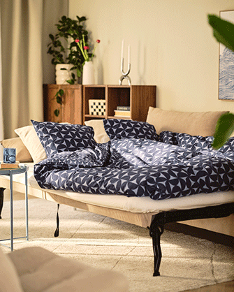 Detalle de un sofá cama abierto con edredón estampado azul y blanco, almohadas y una estantería de madera al fondo.