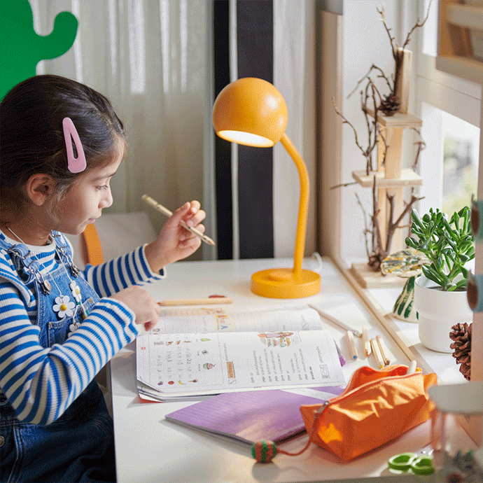 A young girl standing at a white desk by a window, organizing craft supplies and paint next to a white swivel desk chair.