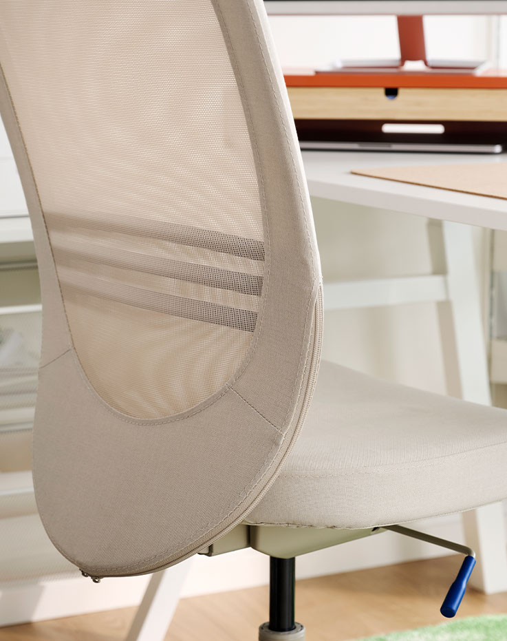 A close-up of a beige FLINTAN office chair with a mesh backrest sits in a bright office space near a desk.