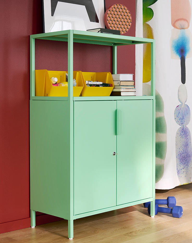 A bright room features a light green TROTTEN cabinet with doors, holding supplies and books beside colourful wall art.