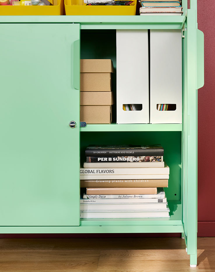 A home office with a light green TROTTEN cabinet with doors, displaying boxes and books on shelves inside.