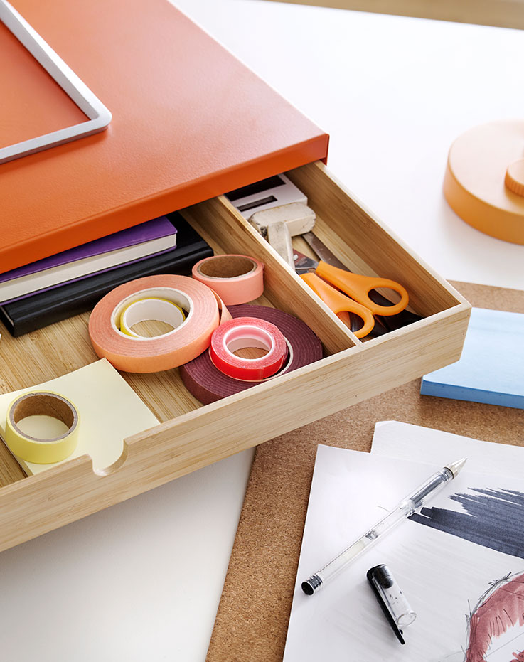 A close-up of a bright orange ELLOVEN monitor stand with drawer, holding stationery items on a desk.