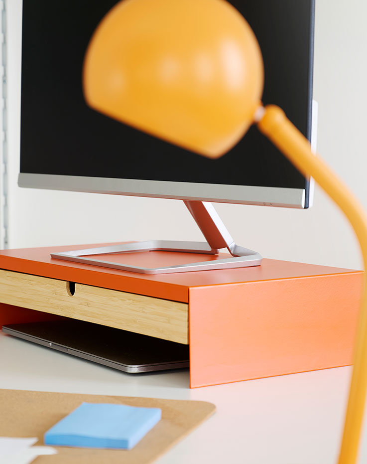A close-up view of a bright orange ELLOVEN monitor stand with a drawer, placed on a desk next to a computer.