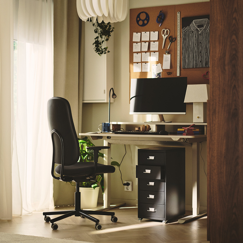 A home office with a beige TRAVSPORT desk sit/stand next to a black rolling chair and potted plants.